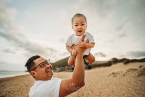 A father lifts his baby boy into the air on a beach at sunset