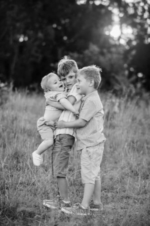 2 boys cuddle their baby brother as they stand in long grass