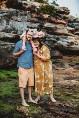 Family Photographer - parents stand on Balmoral beach with their little girl sitting on dad's shoulders