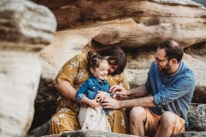 Family Photographer - Parents tickle their little girl sitting on beach rocks