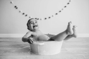 A baby boy laughs, leaning back in a white bath tub after cake smash