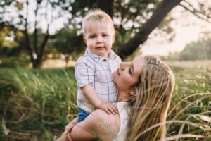 Family Photographer - A mum holds her son in her lap in a grass field at sunset