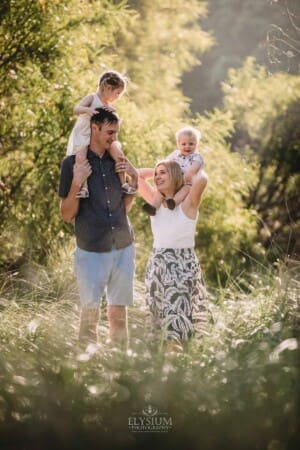 Family Photographer - Parents stand in long grass with their children on their shoulders