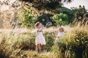Family Photographer - children stand in long grass blowing bubbles at sunset