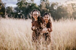 Family Photographer - sisters stand in long grass and giggle while hugging