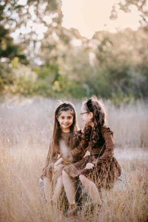 Family Photographer - Sisters hug each other sitting in a grassy field