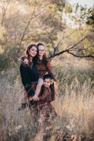 Family Photographer - a mother hugs her 2 daughter standing in long grass