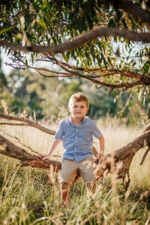 A little boy sits in a curved tree branch as the sun sets behind him