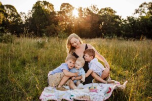 A family sit on a blanket in a grassy field, mother cuddles her 3 boys