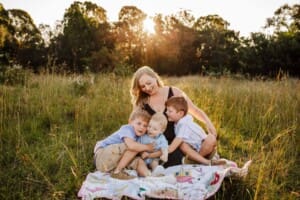 A family sit on a blanket in a grassy field, mother cuddles her 3 boys