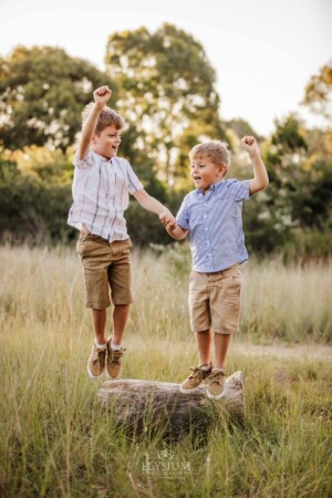 2 brothers jump off a log together in a grass field at sunset