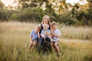 A mother sits on a log, cuddling her 3 little boys