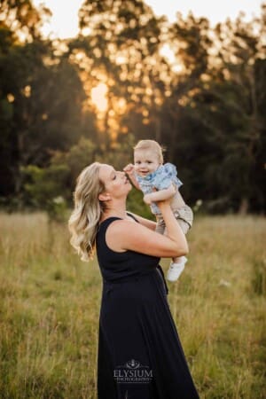 A mother kisses her baby as they stand in a sunny grass field