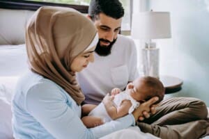 Newborn session - parents hold their baby girl in their arms, sitting on their bed