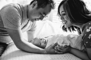 Newborn Session - parents hold their baby between them, laying on a bed