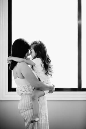 Family session - a mum hugs her little girl, standing in window light