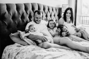 Family session - parents sit with their children on the bed, looking at the baby