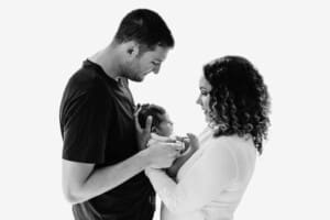 Newborn session - parents stand in front of a window holding their baby between them