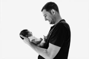 Newborn session - a dad stands against a bright window holding his baby boy