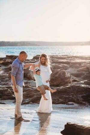 Maternity session - parents stand on a beach and swing their sons feet in the air