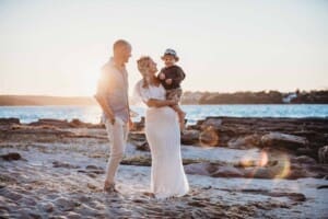 Maternity session - parents hold their boy standing on a beach at sunset