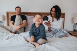 Newborn session - a little boy sits on a bed smiling, his parents sit behind holding the baby