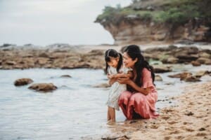 Family Photographer - a little girl stands on the beach cuddling her mum