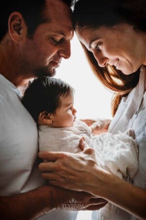 Parents hold their newborn baby girl between them as they stand in front of a bright window