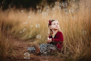 A little girl sits in long grass and blows bubbles