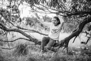 A girl sits in the low branches of a tree at sunset