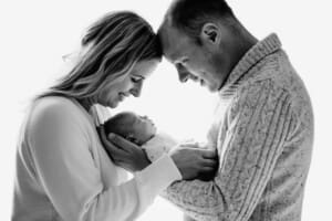Parents hold their newborn baby between them against a bright window