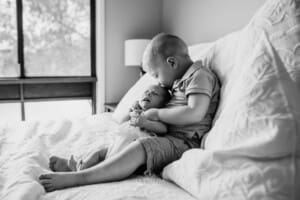 A little boy sits on a bed and kisses his baby sister's head