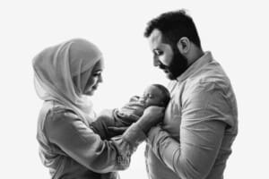 Parents stand against a bright window holding their newborn between them