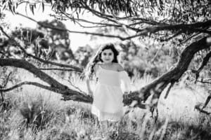 A little girl wearing a white linen dress sits on a low tree branch