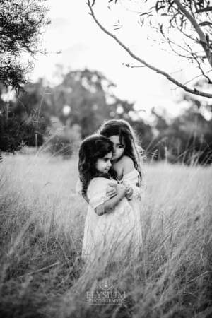 2 little girls in white dresses stand hugging in long grass