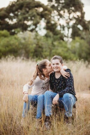 A girl kisses her sister on the cheek as they sit on a log