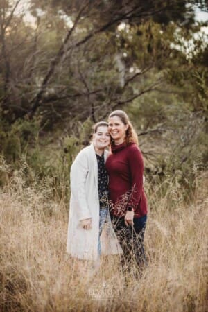 A mother cuddles her daughter as they stand in a grassy field