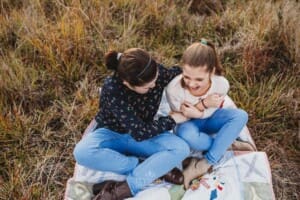 Sisters sit on a rug in long grass and tickle each other