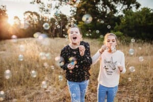 2 girls stand in long grass blowing bubbles at sunset