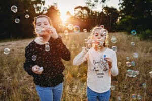 2 girls stand in long grass blowing bubbles at sunset