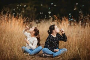 2 girls sit in long grass blowing bubbles at sunset