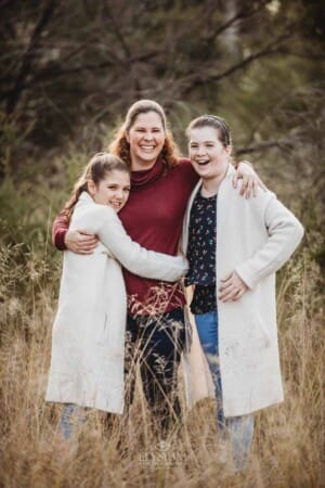 A mother cuddles her daughters standing in long grass at sunset