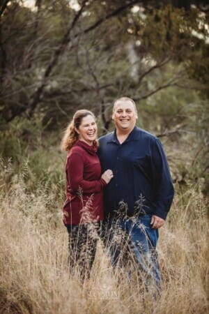 Parents cuddle each other, standing in long grass at sunset