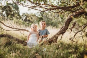 2 children sit on a low tree branch in a grassy field