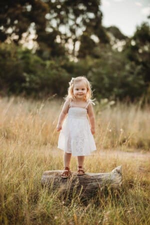 A little girl stands on a log in long grass at sunset