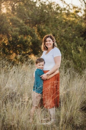 A little boys stands in long grass hugging his mother
