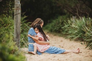 A mother sits cuddling her daughter on a sandy beach path