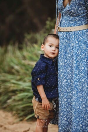 A little boy stands cuddling his mother's leg