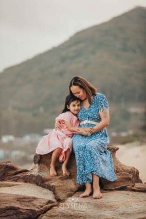 A mother sits cuddling her daughter on rocks at the beach