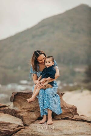 A mother sits cuddling her son on rocks at the beach